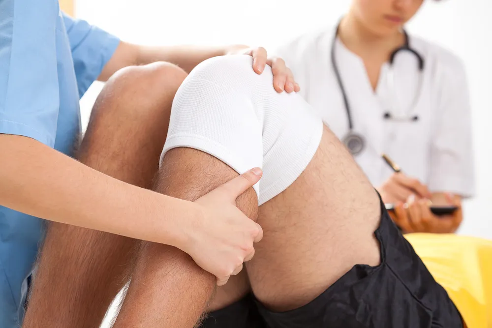 A medical professional examines a patients bandaged knee while another doctor takes notes in the background. The scene captures Injury Recovery at New Life Rehab Clinic, with focus on the patient’s injured, wrapped knee.