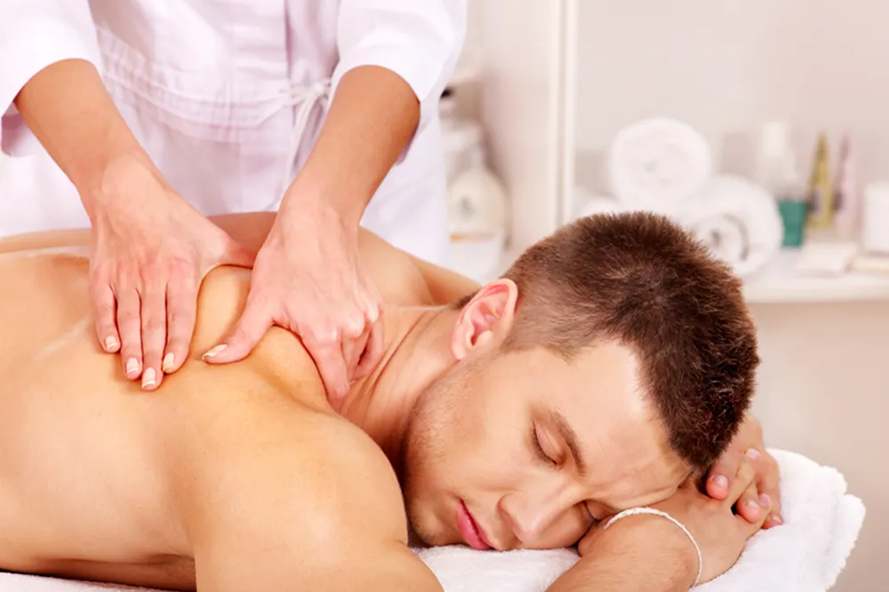 A man lies face down on a massage table with his eyes closed, while a therapist at New Life Rehab Clinic provides expert massage therapy to his upper back and shoulders. Towels and spa supplies are visible in the background.
