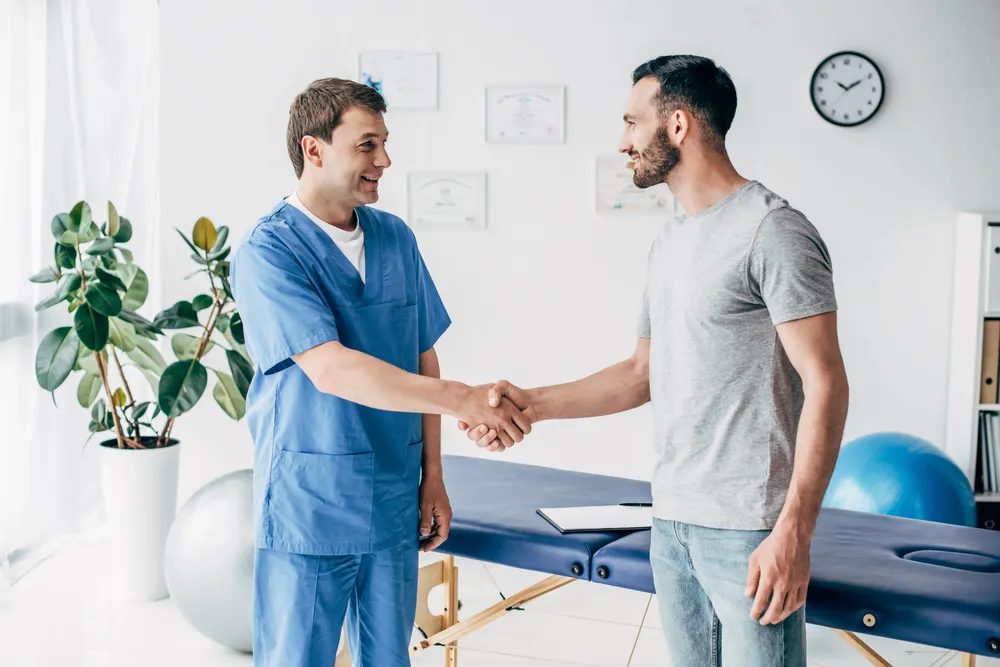 A man in blue medical scrubs and a man in casual clothes shake hands and smile in a bright clinic room with a treatment table, exercise balls, and a clock on the wall.