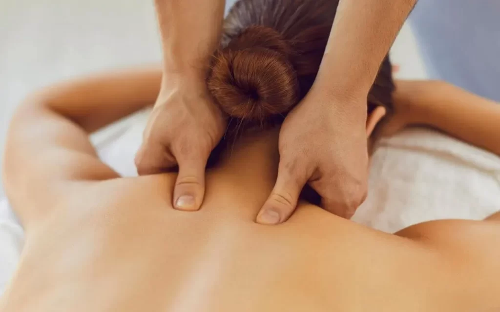 A person with their hair in a bun is lying face down, receiving a back massage. Two hands are pressing on their upper back muscles, suggesting a relaxing massage therapy session.