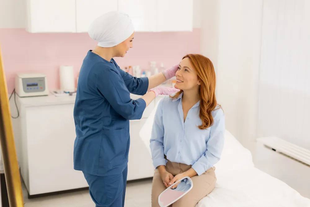 A healthcare professional in blue scrubs and a white head covering talks to a smiling woman with red hair seated on a medical bed, holding a mirror, highlighting Personalized Treatment at New Life Rehab Clinic in a bright clinic room.